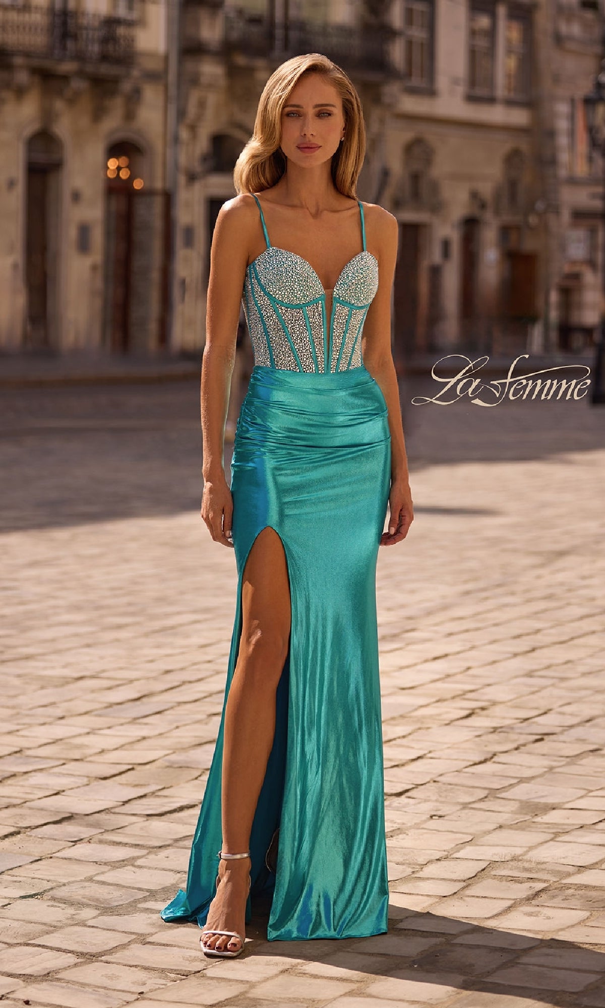 Woman wearing La Femme 32732, a long aqua metallic prom dress with a beaded corset-style bodice, spaghetti straps, and thigh-high slit, paired with silver heels on a cobblestone street.